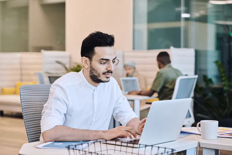 A man in a white shirt typing on a laptop in a bright shared office