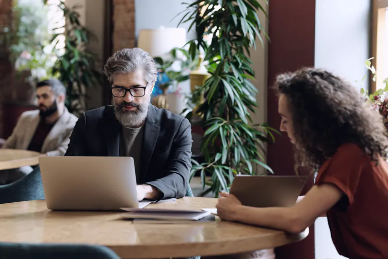 An older man in a dark blazer working on a laptop at a round table opposite a colleague
