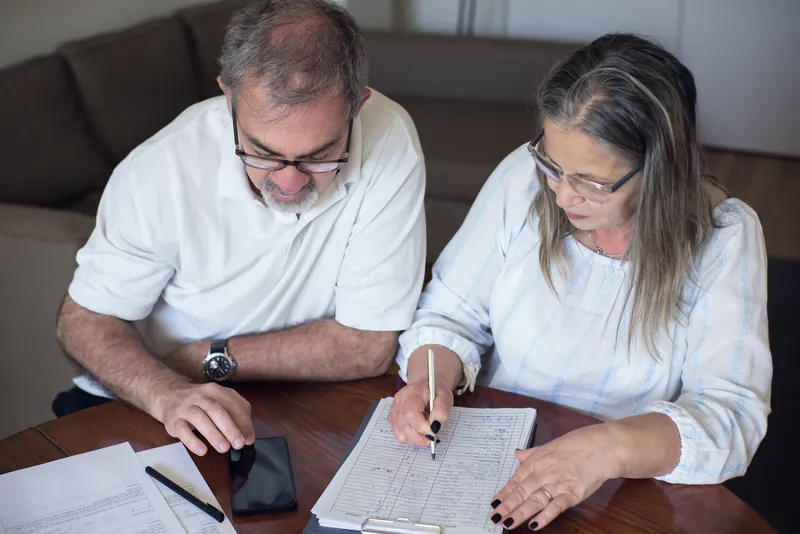 An older couple at a wooden table going through papers together, one of them writing