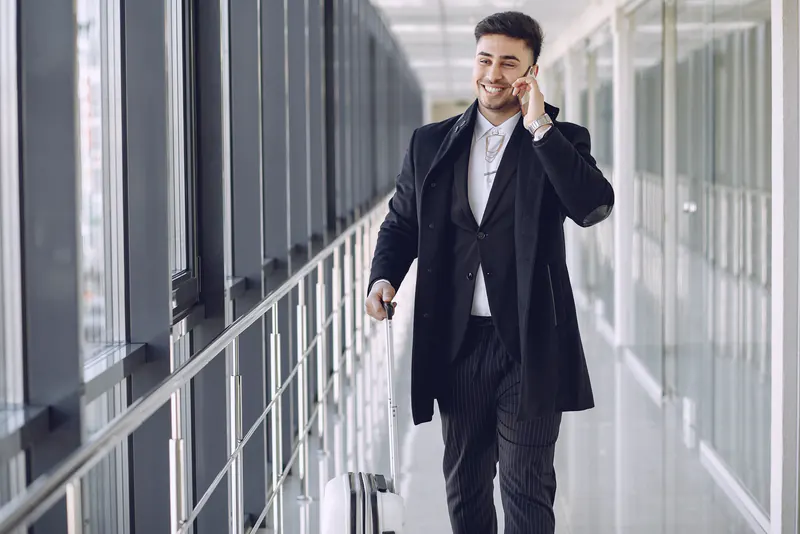 A man in a dark overcoat on the phone, walking through a glass corridor with a suitcase