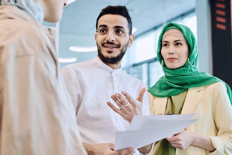 Two colleagues in an office talking over a printed document, one of them wearing a green headscarf
