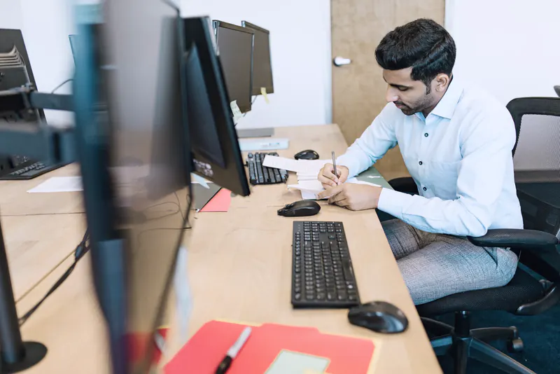 A man in a light blue shirt writing notes at an office desk beside two computer screens