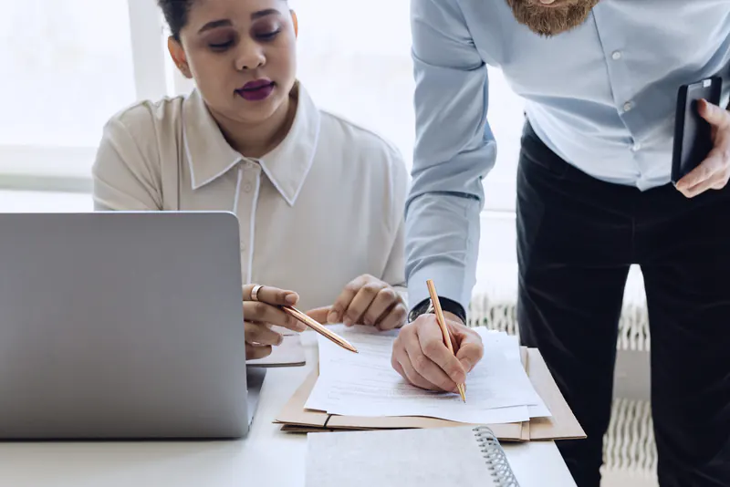 A woman at a laptop working through papers while a colleague leans in to sign a page