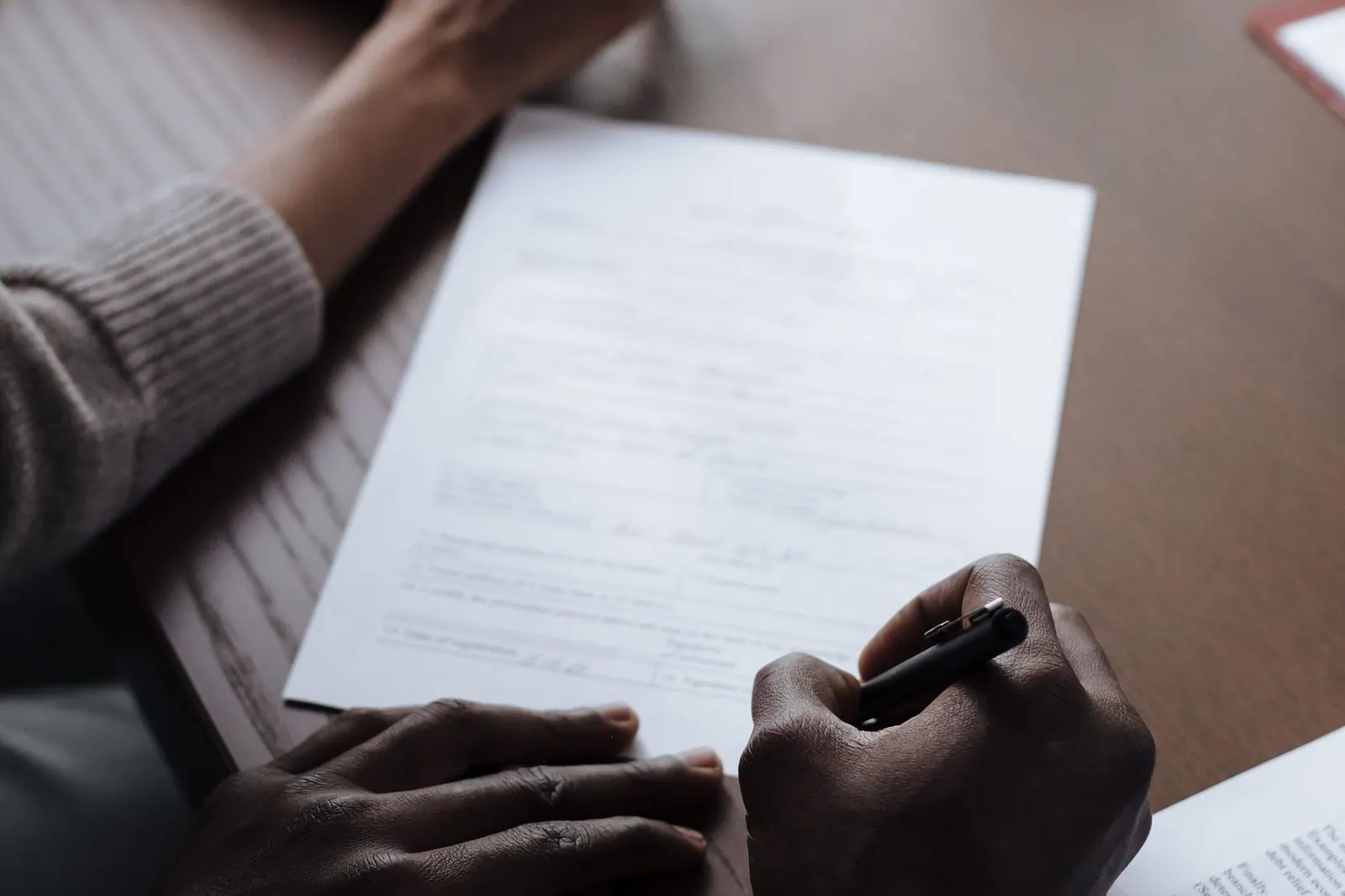 Two people across a wooden table, one signing a printed page with a pen