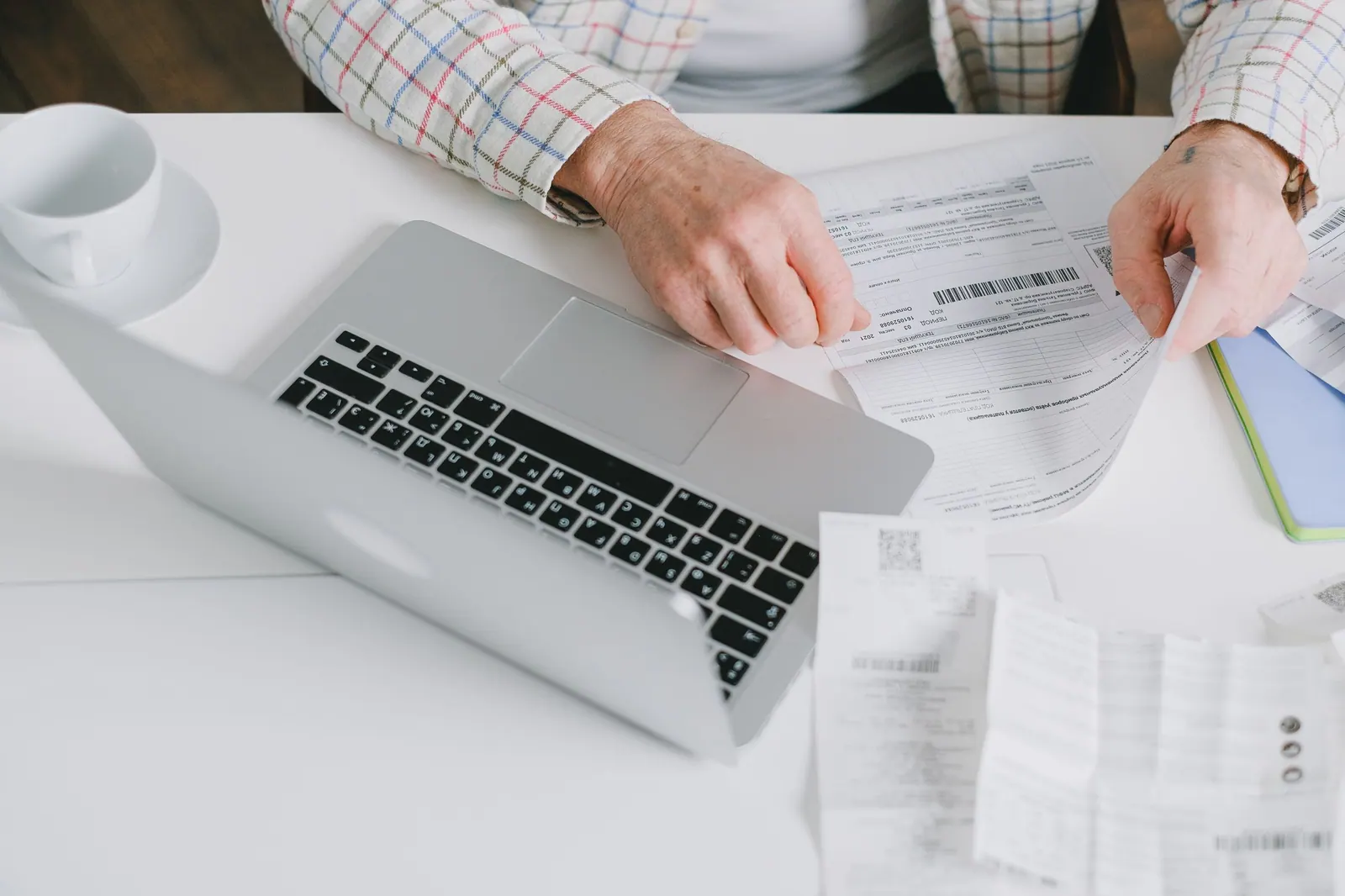 Hands checking a printed invoice against a laptop, more invoices spread on the desk